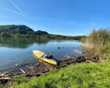 Gelbes SUP-Board liegt am Ufer bereit für SUP Touren Bayern. Ruhiger See, bewaldete Hügel und blauer Himmel.