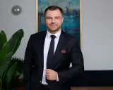 Mężczyzna w garniturze, stojący obok rośliny, z obrazem w tle.