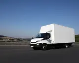 Biały samochód ciężarowy powyżej 3,5 tony pędzi autostradą. W tle krajobraz z pagórkami i domami.