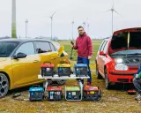 Dwóch mężczyzn testuje, jak samochód jako agregat prądotwórczy zasila sprzęt. W tle wiatraki.