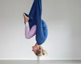 Kvinna hänger upp och ner i en blå tyghängmatta, utövar flying yoga.