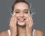 Mujer sonriente con espuma y agua en la cara, disfrutando de su rutina de doble limpieza.