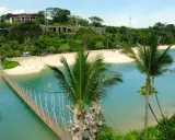 Hängende Brücke über türkisfarbenem Wasser führt zu einem sonnigen singapur strand mit Palmen und Resorts im Hintergrund.