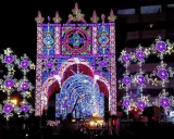 Un tunnel di luci colorate e decorazioni luminose, tipiche delle feste patronali, crea un'atmosfera magica e festosa.