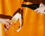 Schaumiges Sektglas läuft über, während eine Flasche auf orangefarbenem Hintergrund eingegossen wird. Eine Szene, die an die **zero dosage definition** erinnert, wo jeder Tropfen zählt.