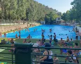 Gente disfrutando de un día soleado en la **piscina segura de la sierra**. Niños y adultos se refrescan en el agua azul, rodeados de árboles y un paisaje montañoso.