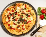 Pyszna pizza z serka wiejskiego, pomidorkami koktajlowymi i bazylią. Obok leżą świeże pomidory i krajalnica do pizzy.
