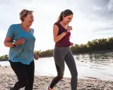 Zwei Frauen joggen am Wasser, um ihre Fettverbrennung anzuregen. Sie lächeln und genießen die Bewegung.