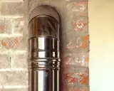 Błyszcząca rura stalowa podłączona do ceramicznego komina, widoczna na tle ceglanej ściany.