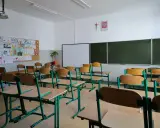 Pusta klasa z rzędami ławek i krzeseł, tablicą i dziecięcymi rysunkami na ścianie. Brak ucznia w tej sali może sugerować nieklasyfikowanie ucznia.