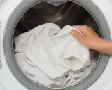 Ręka wkłada białą pościel do bębna suszarki. Szybkie suszenie pościeli w suszarce bębnowej to wygoda.
