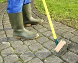 Person en bottes vertes brossant la mousse sur une terrasse pavée. Astuce pour savoir comment enlever la mousse sur une terrasse.