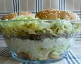 Warstwowa sałatka big mac w szklanej misie, z bułkami na wierzchu.