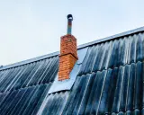 Stary, ceglany komin z metalową rurą wystaje ponad falistą, ciemną blachę dachową. Blacha wokół komina jest przycięta i zabezpieczona.
