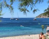 En man sitter på en stenstrand vid havet. I vattnet simmar en person och flera båtar ligger för ankar. En grön kulle reser sig i bakgrunden. En idyllisk vy från Vis, Kroatien.