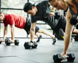 Drei Personen machen eine Plank-Übung mit Kurzhanteln. Ein Mann in Rot, ein Mann mit Kappe und eine Frau in Grau trainieren. CrossFit gesund und stark.