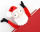 Marcador de libro amigurumi de Papá Noel, con brazos rojos y gorro festivo, asomando de un libro rojo.