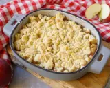 Ein goldbrauner Apfel Crumble in einer Auflaufform, bereit zum Genießen. Daneben liegen frische Äpfel auf kariertem Tuch.
