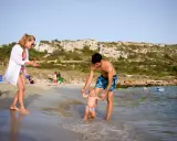 Familia disfrutando de un día en la playa en Menorca con niños. El padre ayuda a su bebé a dar sus primeros pasos en el agua, mientras la madre observa sonriente.
