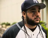 Mężczyzna w czapce z daszkiem i w bluzie z kapturem, z tatuażami na twarzy i szyi, przypominający a$ap yams.