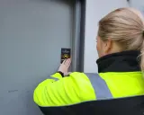 En person med reflexväst fäster en skylt med texten "Kameraövervakning" på en dörr, vilket indikerar ett larm på en byggarbetsplats.
