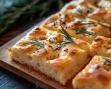 En gyllene focaccia långpanna med rosmarin och kryddor, skuren i bitar på en träbricka.