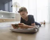 Un adolescent, absorbé par son téléphone, illustre la dépendance aux écrans. Il est allongé sur un coussin, le regard fixé sur l'appareil.
