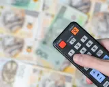 Dłoń trzyma pilota na tle polskich banknotów. Czy abonament RTV i zameldowanie mają związek z kosztami?