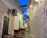 Un vicolo suggestivo nel Val d'Itria, illuminato da lucine, con sedute colorate e muri in pietra bianca.