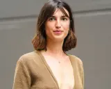 Mujer con cabello castaño ondulado, en transición de pelo corto a largo, luce un suéter marrón y un collar con cruz.