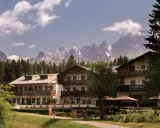 Un hotel alpino immerso nel verde, con imponenti vette innevate sullo sfondo, un perfetto scenario da montagna d'estate.