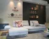 Divano componibile grigio chiaro con cuscini decorativi, logo Calia Italia e "Vogue Xperience" sulla parete.