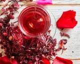 Czerwona, aromatyczna herbata z hibiskusem w szklance z zaparzaczem w kształcie serca, otoczona suszonymi kwiatami i płatkami róż.