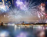 Fyrverkerier lyser upp himlen över Stockholm vid nyår. Vatten reflekterar ljusen från staden och fyrverkerierna.