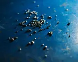 Piccole perle di argento riciclato sparse su una superficie blu scuro, con riflessi luminosi.