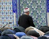 Männer in einer Moschee, die sich im Gebet verneigen. Der Imam trägt einen weißen und roten Hut. Die Fliesenwand ist mit islamischer Kalligrafie und Mustern verziert. Dies ist ein Ort, an dem der größte Sünde im Islam, Shirk, vermieden wird.