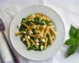 Pyszny makaron penne ze szpinakiem i twarogiem, udekorowany listkami bazylii. Obok leży widelec i biała serwetka.