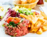 Un délicieux tartare de boeuf, garni de persil et d'une tranche de tomate, accompagné de frites dorées et d'une salade fraîche.