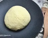 Ein runder, heller pizzateig italienischer Art liegt auf einem schwarzen Teller. Daneben ein Holzspatel und ein Küchentuch.