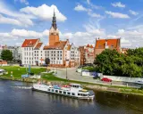 Statek wycieczkowy na rzece przy nabrzeżu z kamienicami i wieżą kościelną w tle.