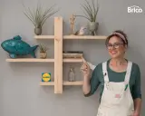 Una mujer sonriente muestra una estantería de madera, decorada con plantas, un pez azul y libros. Ideal para aprender como hacer una estantería de madera.