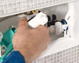 Ręka majstruje przy mechanizmie spłuczki podtynkowej, przygotowując naprawę.
