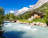 Val Ferret: un'incantevole casa in pietra alpina vicino a un fiume turchese, con un ponte e montagne innevate sullo sfondo.