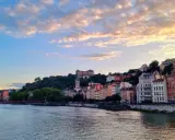 Blick auf die Stadt Lyon, Frankreich, mit dem Fluss Saône im Vordergrund und bunten Gebäuden, die sich den Hügel hinaufziehen. Ein Schloss thront über der Stadt.