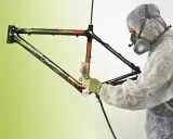 Person in Schutzanzug lackiert kunstvoll einen Rennrad Rahmen mit Spraydose.