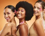 Trois femmes souriantes, un vrai "glow up traduction" avec des fleurs dans les cheveux et un maquillage éclatant.