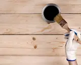 En hand med vit handske doppar en pensel i en burk med svart olja för tryckimpregnerat trä.