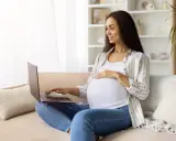Kobieta w ciąży na kanapie szuka w internecie najdokładniejszy termin porodu, uśmiecha się.