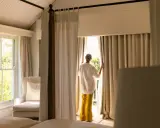 Una mujer abre cortinas para dormitorios, dejando entrar la luz. Se ven camas y un sillón junto a la ventana.