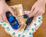 Manos sostienen un coche azul de juguete, llaves y billetes de euro. El coche no arranca, quizás por falta de dinero.